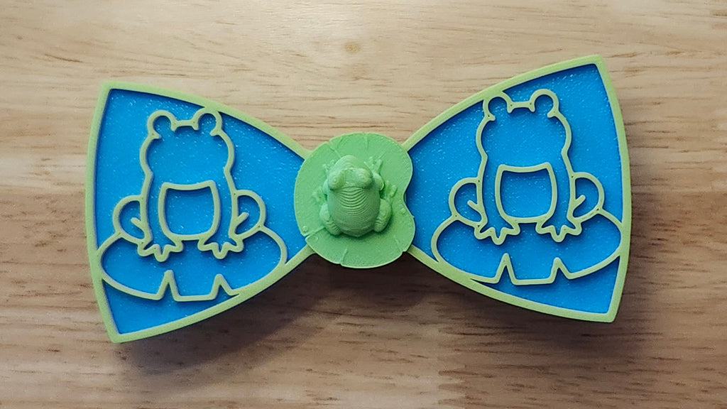 Frog Button Cover with Lily Pad Bow (reverse side shown), Frog Outline Bow (green side). 3D printed for use as a bow tie, button cover, necklace, pin, hat pin, lapel pin and so much more. 