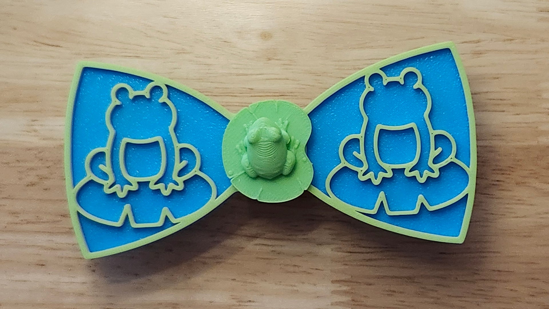 Frog Button Cover with Lily Pad Bow (reverse side shown), Frog Outline Bow (green side). 3D printed for use as a bow tie, button cover, necklace, pin, hat pin, lapel pin and so much more. 