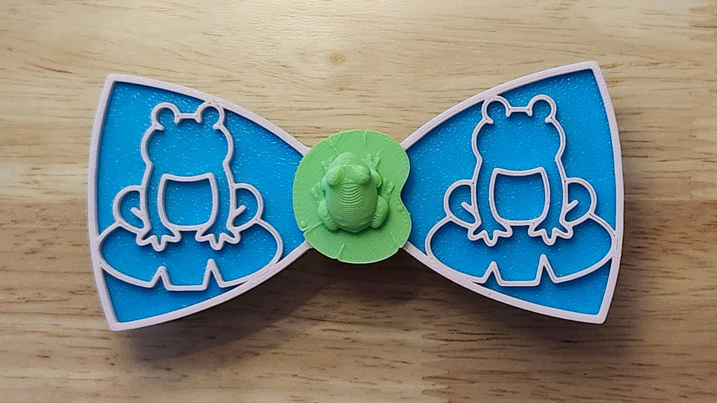 Frog Button Cover with Lily Pad Bow (reverse side shown), Frog Outline Bow (pink side). 3D printed for use as a bow tie, button cover, necklace, pin, hat pin, lapel pin and so much more. 