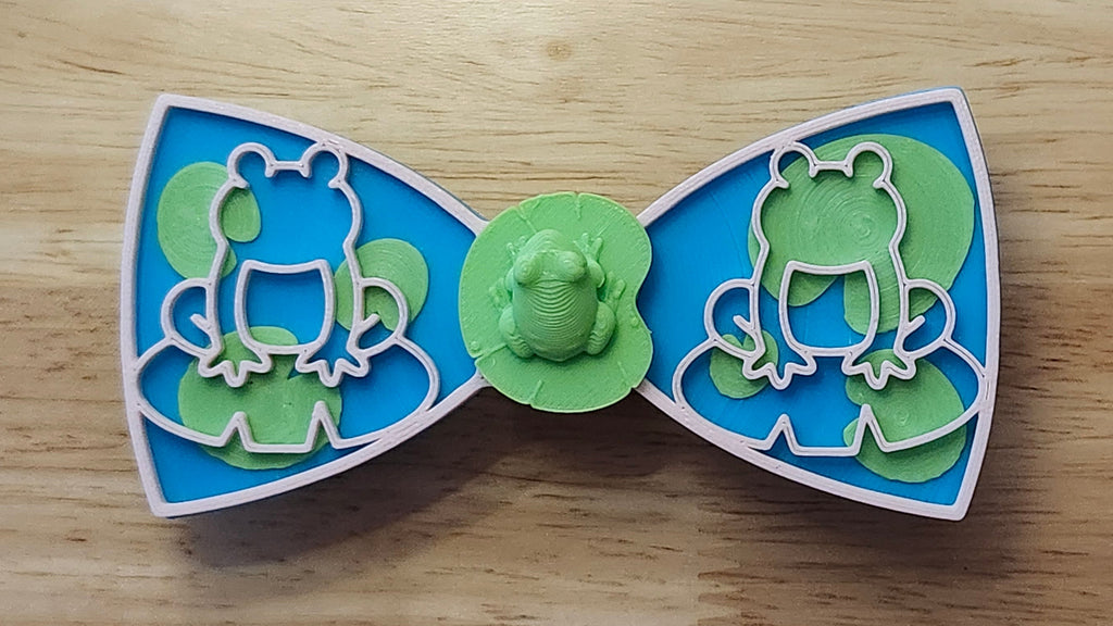 Frog Button Cover with Lily Pad Bow, Frog Outline Bow (pink side). 3D printed for use as a bow tie, button cover, necklace, pin, hat pin, lapel pin and so much more. 
