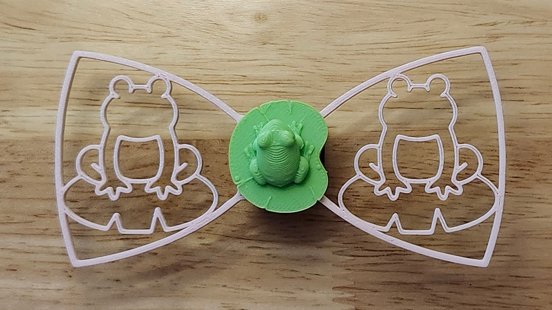 Frog Button Cover with Frog Outline Bow (pink side). 3D printed for use as a bow tie, button cover, necklace, pin, hat pin, lapel pin and so much more. 